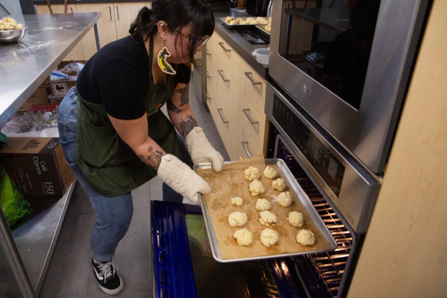 A photo of a younger person wearing jeans, sneakers, a tshirt, sturgeon beaded earrings, an apron and oven mitts pulling a baking sheet with bannock on it out of the oven.