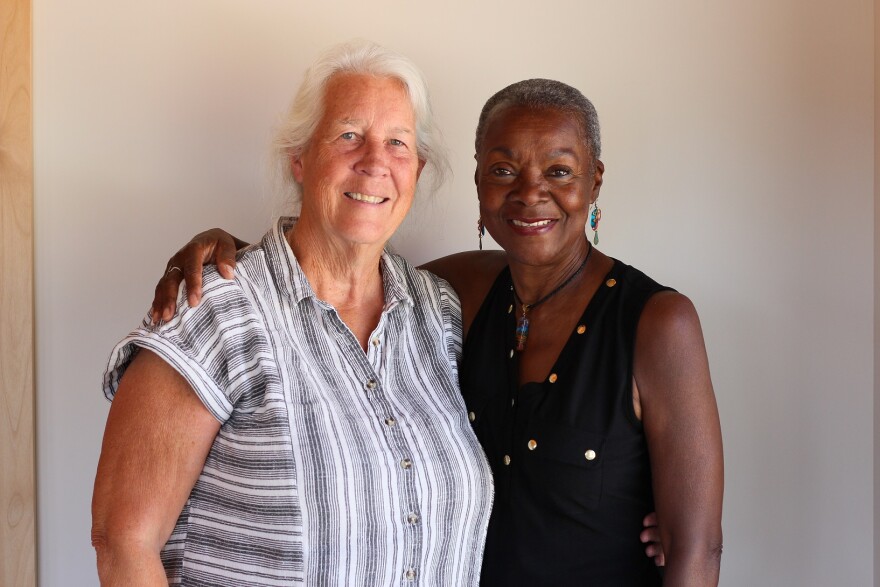 Teri Guy and Janice Brooks at their StoryCorps appointment in Springdale, Utah in May 2025.  Teri  Guy  is a white woman. She has medium length white wavy hair  parted on the side, and she wears a grey and white striped, short-sleeved, button up blouse with a collar.  She has her left arm around the waist of Janice Brooks who stands beside her.  Teri stands a few inches taller than Janice and is wider.  Janice is African American.  She has very short gray hair and wears dangling earrings and a pendant from a choker necklace.  Her top is black and sleeveless with gold button details.  Janice has her right arm around Teri's shoulders.