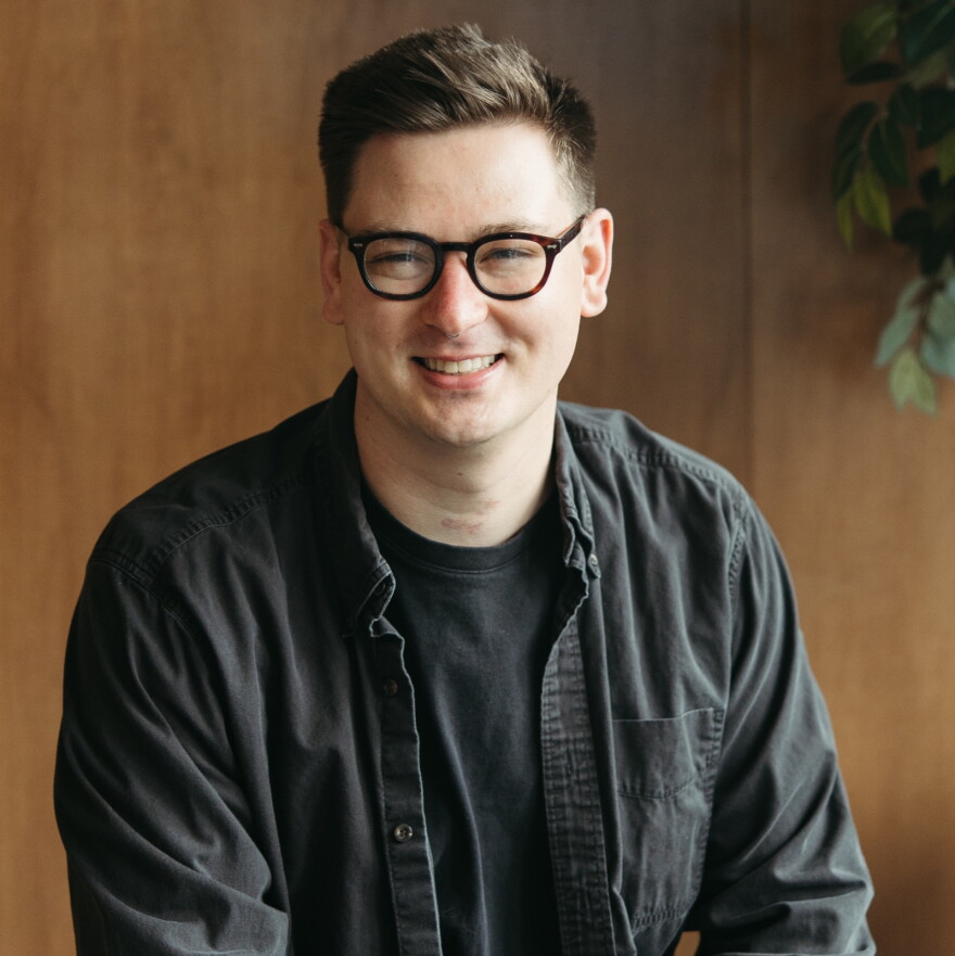 a man in a dark shirt with glasses poses in front of a brown background