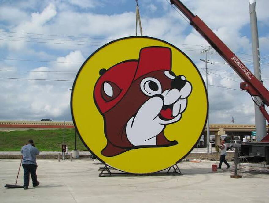A large circular sign showing Buc-ee’s beaver mascot, a smiling cartoon beaver in a red cap with a white-gloved paw raised, on a yellow background with a dark outline. The sign rests on a metal stand on a concrete lot while a crane holds it up from above. Two workers stand nearby, one sweeping and one by a red bucket, with an overpass and cloudy sky behind them.