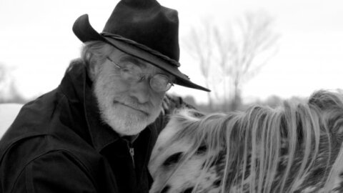 A black and white photo of a man in a cowboy hat next to a horse, with a bare tree against the white sky.