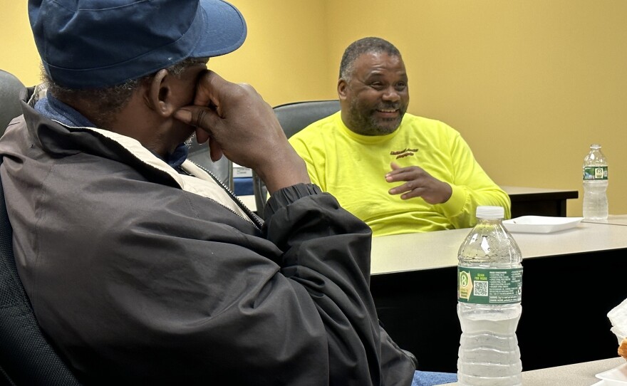 A Black man, turned with his back to the camera, listens to another Black male tell a story as he laughs.
