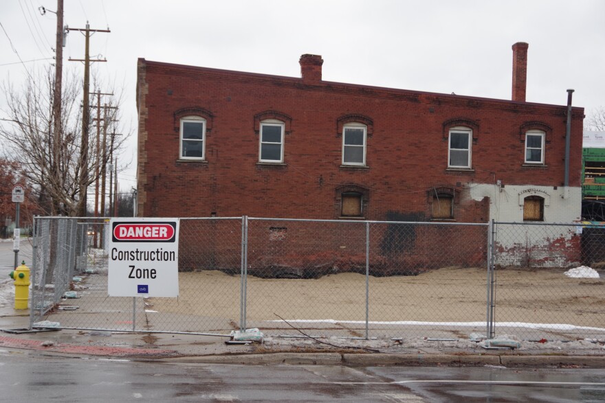 A cold gray chain link fence wraps around a wet patch of sand, a sign declaring the area as a construction zone hangs from the make shift barrier. A dilapidated dark red brick building sits just behind the patch of sand, with light brown ply wood boards a few blocking windows at its base. Small puddles on the adjacent street and gray clouds above denote the rainy weather that occurred that day. 