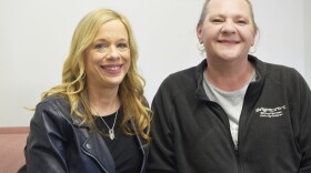 Two women sit together and smile for the camera, each are wearing black and smiling. The woman on the right is slightly taller. 