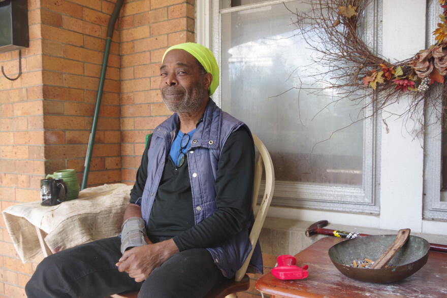 A man in a long black shirt, black loose-fitting pants, a dark blue vest and a bright green beanie sits in a plastic lawn chair in front of the window to a brick house. To the right sits a cane and an ash tray on a wooden table. To the left sits a black mug and a green mug, which are place on a table covered with a white towel.