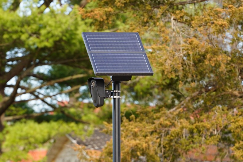 A solar-powered security camera is mounted on a tall black metal pole outdoors. The rectangular solar panel sits angled at the top of the pole, while the small black camera module is attached just below it. Green and brown autumn tree foliage blurs softly in the background.