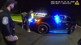 Two men are being handcuffed by a border patrol agent as they stand in front of the hood of a police car. In the foreground, a police officer with a Southgate police emblem on his shoulder watches the scene. It is dark, but a brightly-lit gas station can be partly seen in the background. 