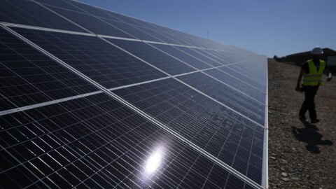 A close up of a ground mounted solar panel. A construction worker in a hard hat and a yellow safety vest walks past the row of panels in the background. 