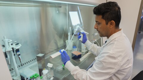 Man in lab coat and blue-gloved hands works at fume hood with vials, pipettes and other scientific equipment