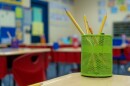 A green pencil container with yellow pencils is in the foreground. Red chairs and blue carpet from an elementary school are blurred in the background