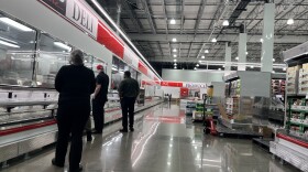 Three Costco employees stand in front of mostly empty deli shelves in a Costco aisle. A forklift sits in the aisle. 