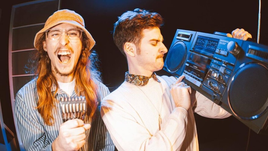  A man with long hair and glasses wearing a bucket hat yells toward the camera as another man next to him looks at a vintage boombox while pressing the play button.