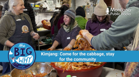 People gather in a cozy, bustling kitchen space at Winter Kissed Farm in Montana’s Bitterroot Valley, preparing kimchi during a traditional Korean kimjang event. In the center, Sohn Jung-a, one of the organizers, smiles while helping mix ingredients in a large metal bowl. Around her, other participants in winter clothing work together, handling napa cabbage coated in red spices. A large tub of kimchi sits in the foreground. A banner reads: "The Big Why – Kimjang: Come for the cabbage, stay for the community."