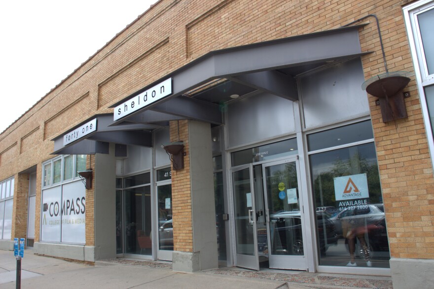 The front of the building at 41 Sheldon in Grand Rapids. The facade shows orange brick. In the window, a realtor sign still shows "Available." In a window in the background is a sign for the Compass College of Film & Media, which previously occupied the building