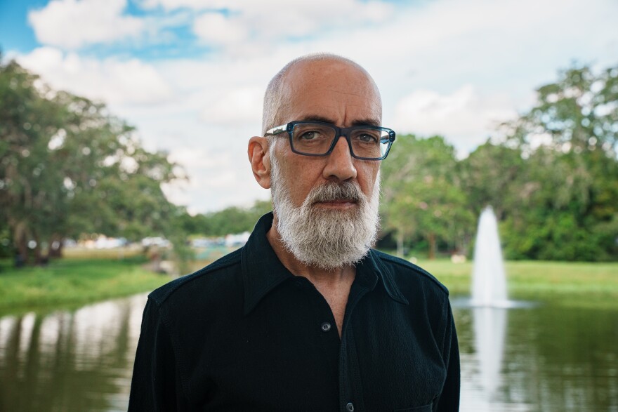 A man with shorn hair, dark glasses with a grey beard and a black shirt stands in front of a fountain. 
