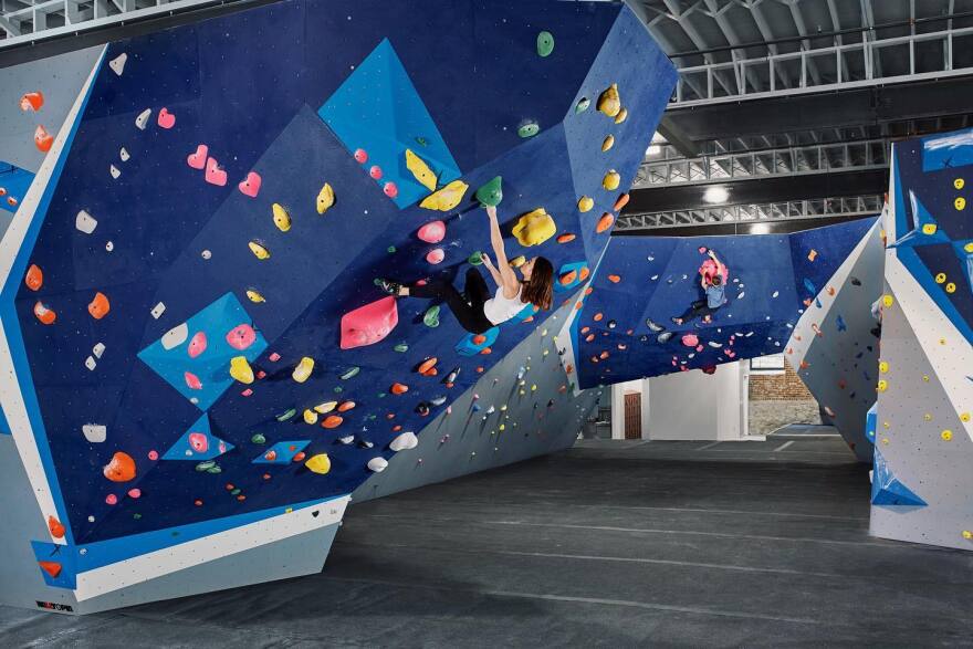 A white woman in a white shirt and black pants grabs on to green handhold on a blue climbing wall. 