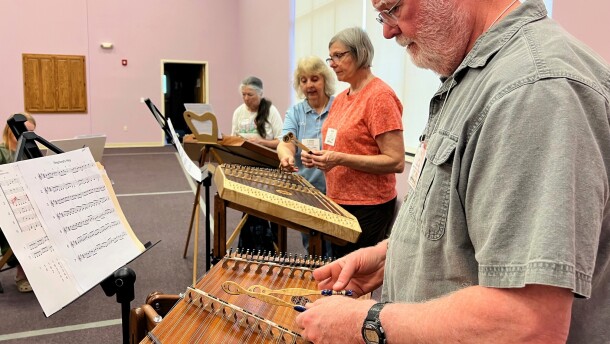 A circle of students stand in front of their hammered dulcimers, a trapezoidal string instrument which they strike with wooden mallets.
