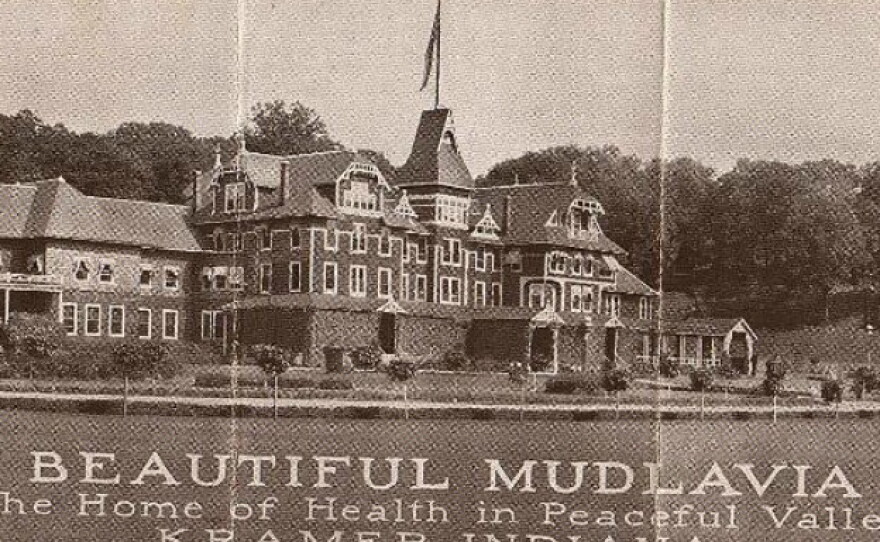 An archival publicity photo showing a large, multi-story Victorian-style hotel building with text at the bottom that reads 'BEAUTIFUL MUDLAVIA, The Home of Health in Peaceful Valley, KRAMER, INDIANA.'