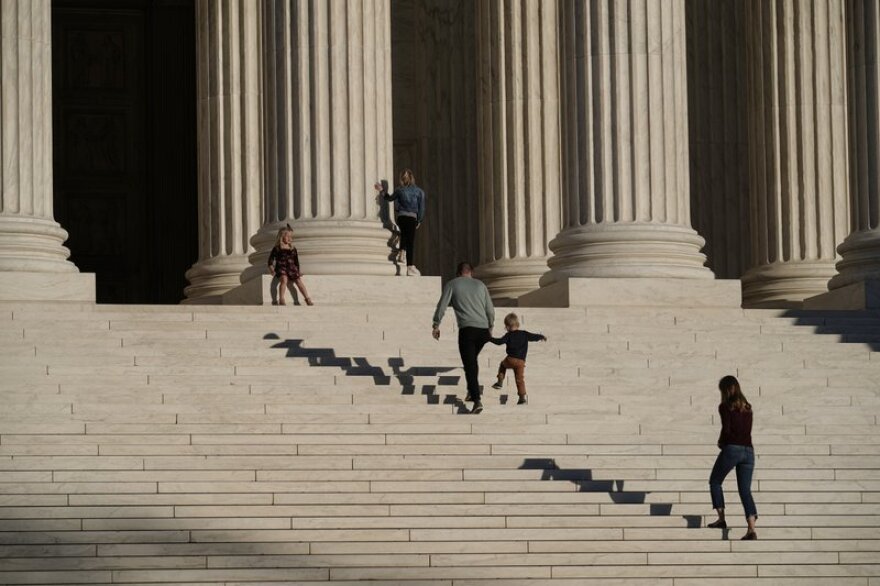 A man holds the hand of a small child as they climb the steps in front of the U.S. Supreme Court Building in Washington, D.C.  A woman follows behind. Their shadows ripple on the steps in front of them.