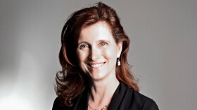 A smiling woman with long brown hair wears a black shirt and jeans.