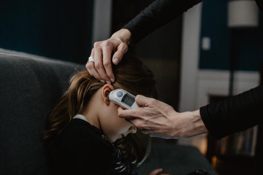 A young girl is seated looking downward, adult hands touch the child's head an hold a thermometer to her ear.jpg