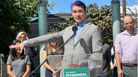 A man in a suit with purple hair stands in front of a transparent podium with an San Francisco Environment Department placard. He gestures to the group of people standing behind him.