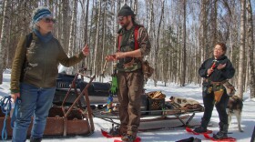 Three people in snowshoes stand around a snow machine in a birch forest