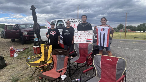 Four people hold protest signs and flags in front of a truck with more signs on it. Lawn chairs are set up in front of them.