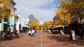 A brown brick road leads forward towards a white structure. Buildings line the path and several groups of people walk in the area. It's a bright day, and the sky is mostly white with clouds, and yellow trees line the path on both sides.