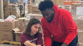 A young woman with brown hair is wearing a burgandy shirt. She has multi-colored plastic Easter eggs in front of her. It appears she is stuffing them with candy as a man earing a red sweatshirt is standing behind her and helping her.