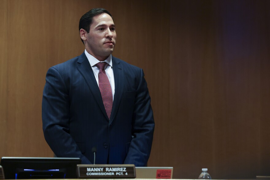 A photo of Tarrant County Commissioner Manny Ramirez, standing up at a dais wearing a blue suit and smiling slightly.