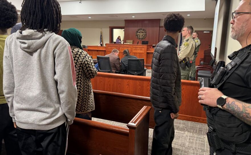 View from the back of a courtroom. Everyone is standing, except for two men at a table in the background. There are law officers to the right. 