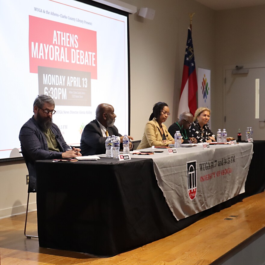Candidates sit at a table during an Athens mayoral debate, with an event slide projected on a screen behind them.