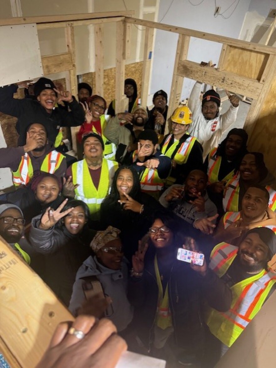 A group of trainees in a CNY ONRAMP construction program wearing yellow safety vests, standing inside a small room.  