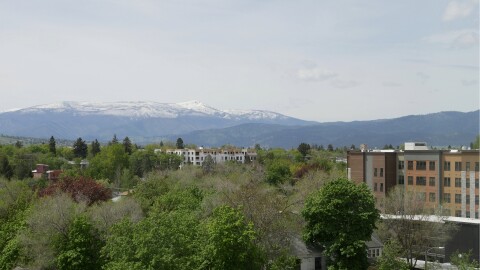 A wide view of Missoula, Montana, shows leafy green neighborhoods and low-rise buildings beneath snow-capped mountains under a hazy spring sky.