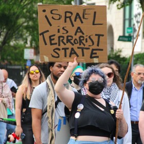 A group of protesters carry sign and Palestinian flags. A woman carries a sign that reads, "Israel is a terrorist state." 