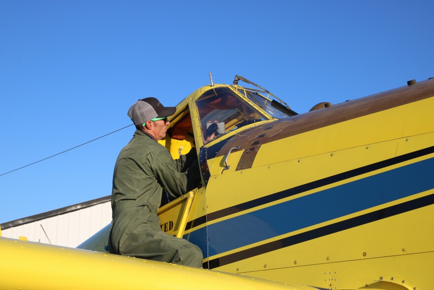 A man in a green jumpsuit, sunglasses and a ballcap looks into the cockpit of a yellow airplane with a blue and black stripe down the side.