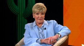 In a screenshot, Betsy Price sits with an arm over a chair, wearing a light blue suit, giving her final state of the city address.