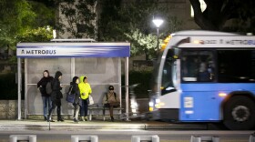 A Capitol Metro bus approaches a group of people as they wait at a CapMetro bus station on Guadalupe Street at the University of Texas. They're wearing jackets because it's cold outside.