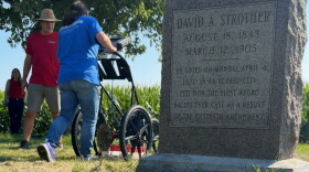 a memorial gravestone is in the foreground. In the background, a volunteering pushes an underground radar device while a man in a red t shirt watches