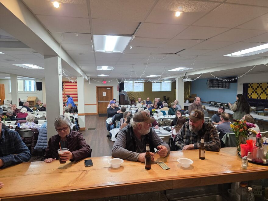 A large legion hall with people sitting at tables eating a Thankgiving buffet and a couple guys in front sitting at a bar drinking beer 