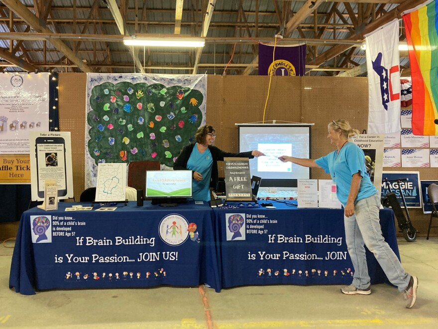 Two woman at a recruiting booth for early childhood educators. Poster of a hand-painted tree hangs in the background.