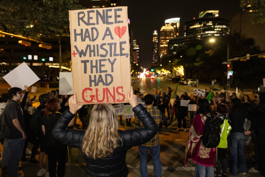 A woman holds a sign reading "Renee had a whistle, they had guns" above her head. She is in the foreground and a crowd and downtown street scene is seen in the background. 