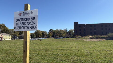 in the foreground, a white sign on a wooden post. in the background, an open field and brown brick building