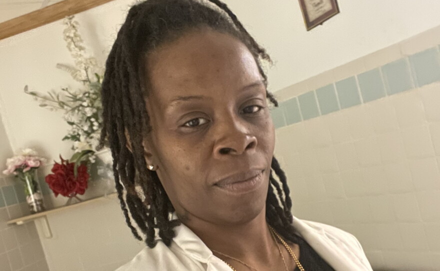 A smiling woman with short black braids wearing gold necklaces and a black top under a white blazer