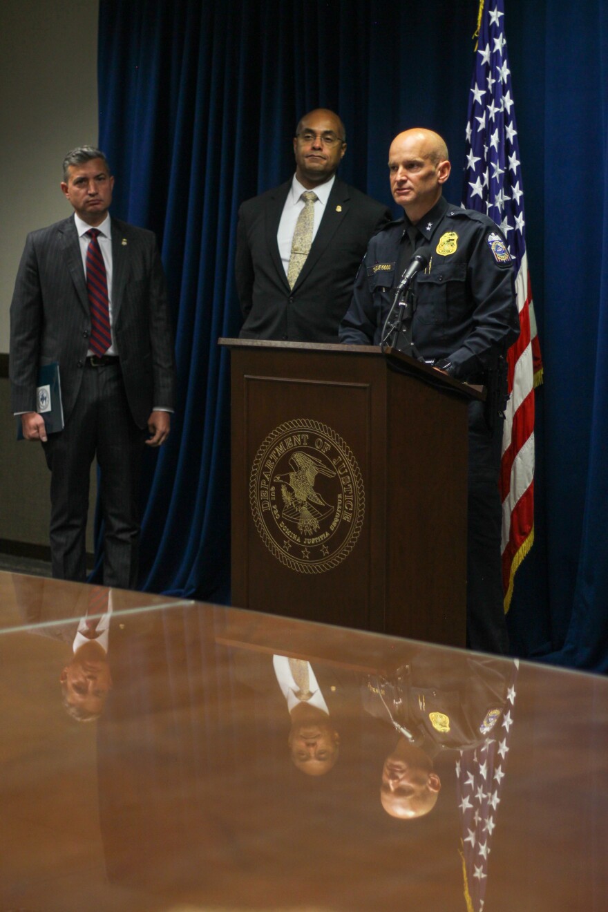 Two men in suits look on as a uniform police officer speaks at a podium flanked by two flags. 
