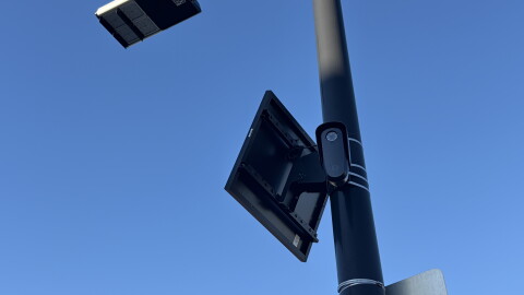 A security camera is affixed to a light pole. The photo is a worm's eye view. The camera has a very small solar panel above it, also affixed to the pole. 