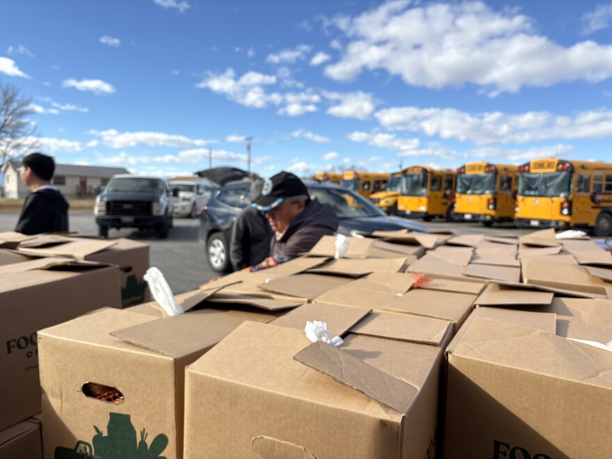 Cardboard boxes with food are stacked in the foreground. Behind them, volunteers help move boxes into a line of cars waiting in a school parking lot.