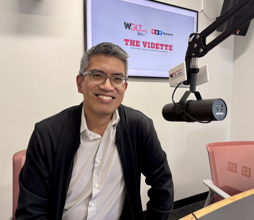 A man wearing glasses and a black jacket smiles while seated in a radio studio. A large microphone and "WGLT" branding are visible, along with a screen displaying "THE VIDETTE.