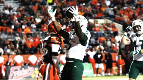 football player in white jersey #5 white helmet and green pants celebrates with both arms raised, football in one hand